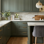 A luxurious kitchen featuring sage green shaker cabinets, white marble countertops with grey veining, and gold hardware. The scene includes a marble kitchen island with grey velvet barstools, brass globe pendant lights, and a bowl of fresh green apples on the counter.